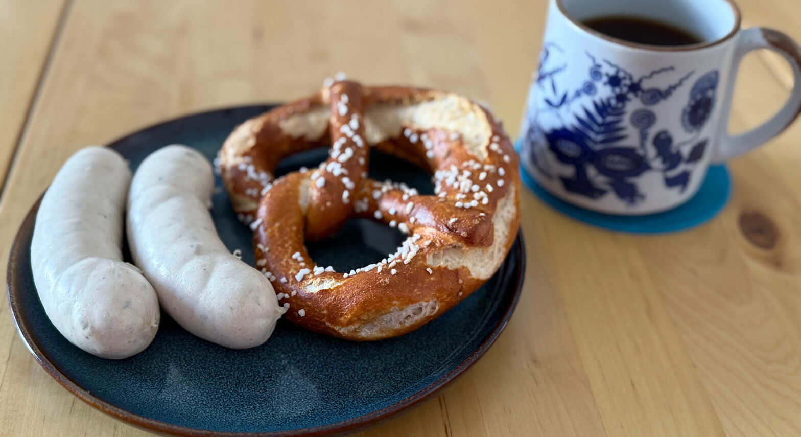 Eine bayerische Breze mit zwei Weisswürsten auf einem blauen teller mit einer Tasse mit blauem Porzellanmuster